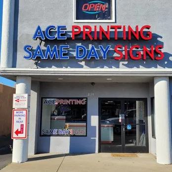 Exterior storefront shot of the Ace Printing / Same Day Signs shop with blue-and-red channel letters above the entrance, a red neon OPEN