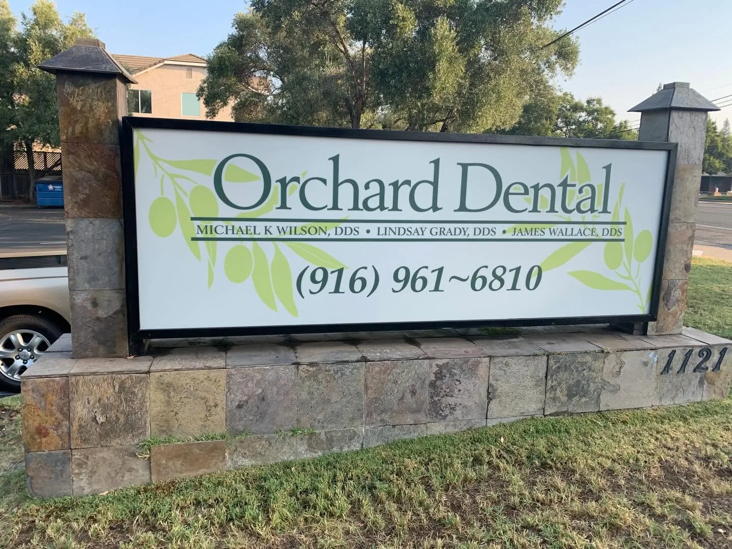 Outdoor monument sign for Orchard Dental with olive-branch artwork mounted in a black frame between stone pillars at street address 1121.