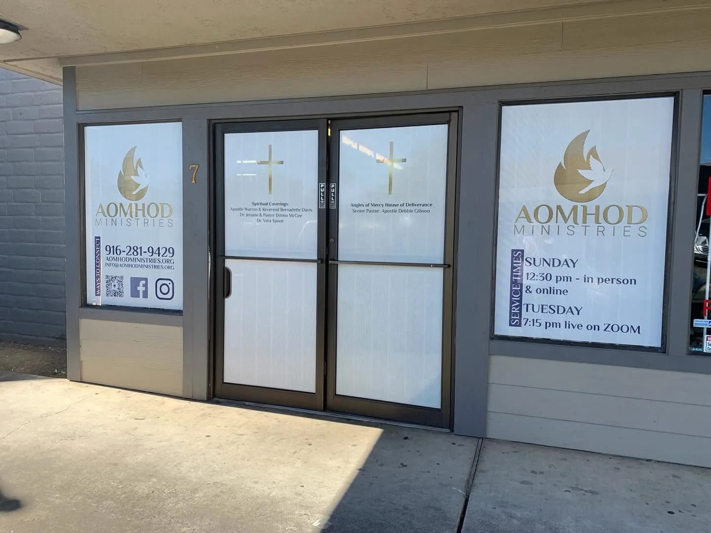 Storefront window and door graphics for AOMHOD Ministries showing gold flame-and-dove logos, service times, contact info, and cross decals