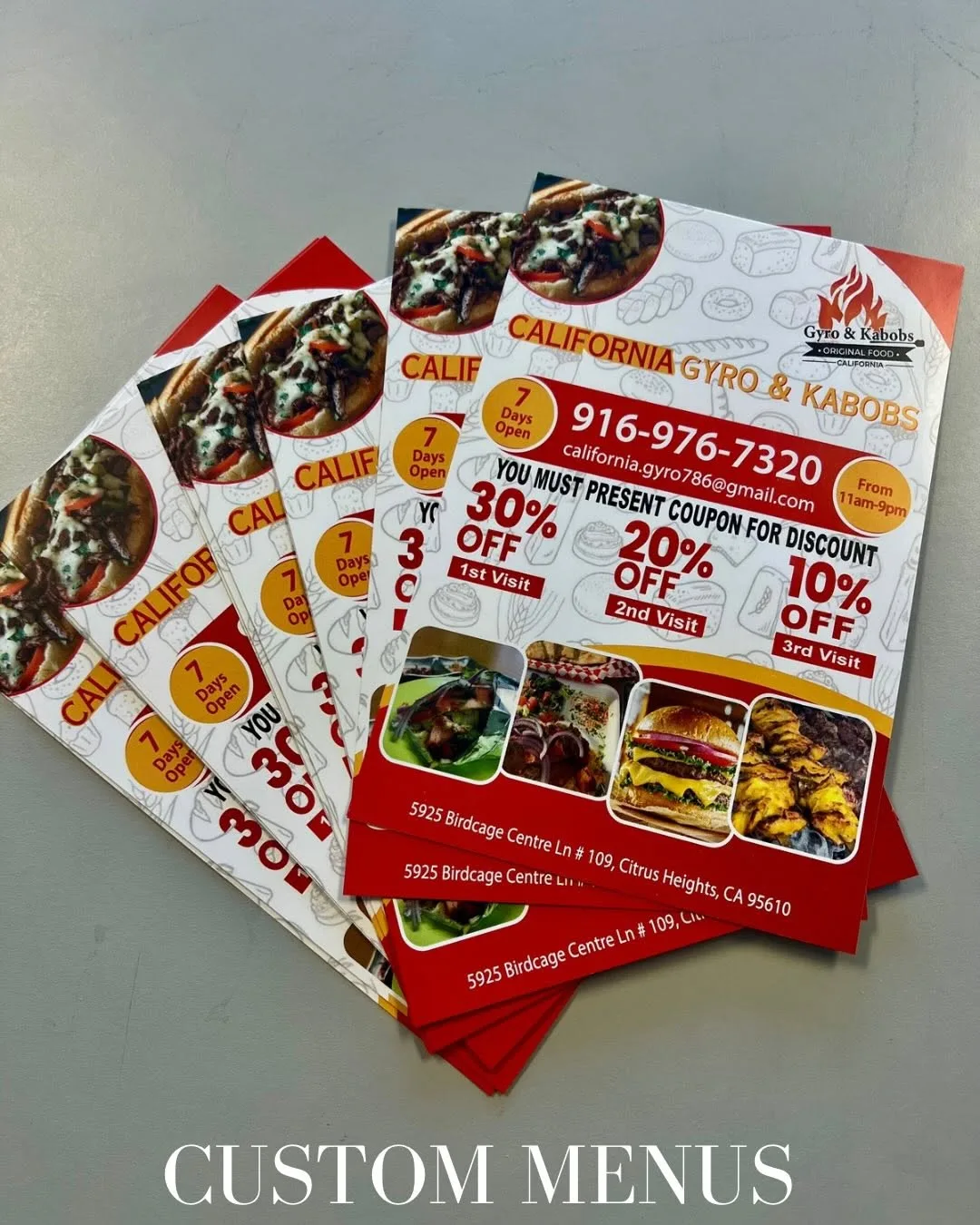 Fanned stack of freshly printed full-color coupon flyers for California Gyro & Kabobs on a work table, with a 'CUSTOM MENUS' text overlay
