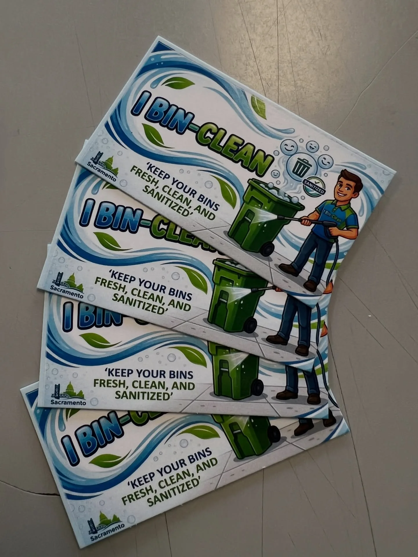 Four printed marketing cards for 'I Bin-Clean' (a Sacramento trash-bin cleaning service) fanned out on a table, featuring a cartoon of a