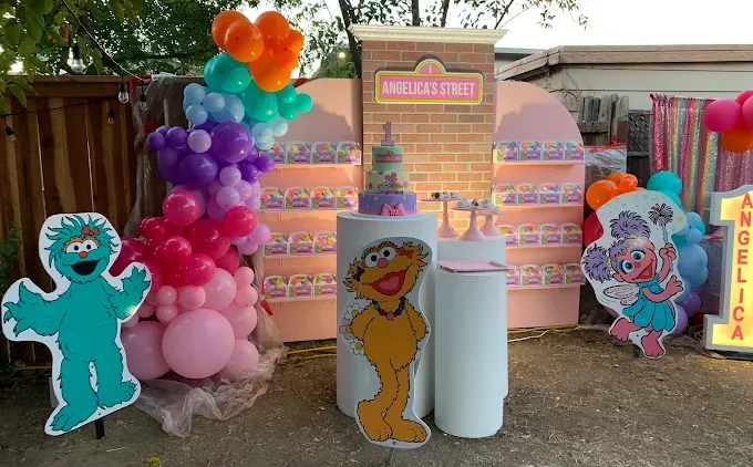 Backyard first-birthday party setup with large printed Sesame Street character cutouts (Rosita, Zoe, Abby Cadabby), a brick-look backdrop