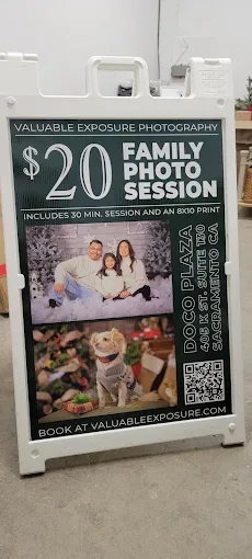 White A-frame sidewalk sign in a warehouse holding a printed poster advertising a $20 family photo session for Valuable Exposure