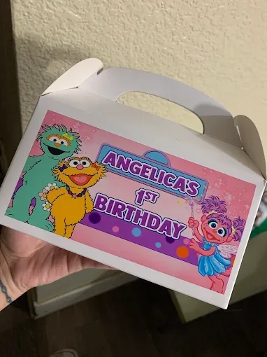 Hand holding a small white gable favor box with a custom-printed Sesame Street themed label reading Angelica's 1st Birthday.